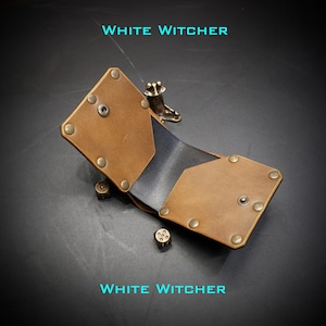 May include: A tan leather dice tray with a black interior, secured by metallic rivets and snaps. The tray features the text WHITE WITCHER in cyan. Two small metallic dice are positioned near the tray, and a metallic frog figurine sits on the edge.