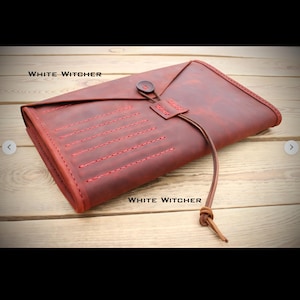 Può includere: Una custodia arrotolabile in pelle rosso scuro con il testo WHITE WITCHER, una chiusura a patta fissata da un bottone e un lungo cordino in pelle, e linee di cucitura rosse decorative sulla superficie anteriore dell'accessorio rettangolare.