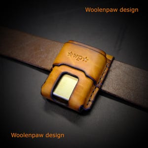 May include: A tan leather belt pouch with a rectangular cutout revealing a metallic gold insert. The pouch features a stamped logo with the text "wp" flanked by two stars. It is threaded onto a brown leather belt. The text "Woolenpaw design" appears.