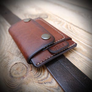 May include: A brown leather wallet with a fold-over flap secured by two metallic snap buttons. The wallet features visible light-colored stitching along the edges and a small rectangular leather tab embossed with the text KRP and a star symbol.