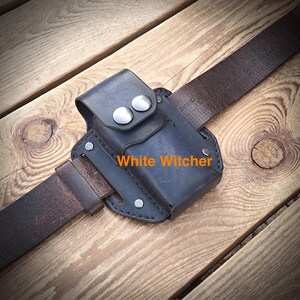 Peut inclure: Une pochette en cuir bleu foncé avec une fermeture à rabat fixée par deux boutons-pression en métal argenté, attachée à une ceinture en cuir marron foncé. Le texte White Witcher est imprimé en orange sur le devant de la pochette à rivets.