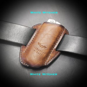 May include: A brown leather belt holster featuring a stamped logo and the text "Enchanted for luck" threaded onto a black leather belt. The holster has visible stitching along the edges and holds a small metallic tool at the top opening.