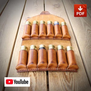 May include: A tan leather shotgun shell holder featuring two rows of six individual stitched slots. Each slot contains a single shotgun shell with a brass base. The leather holder has a pointed top flap and is constructed with visible stitching.