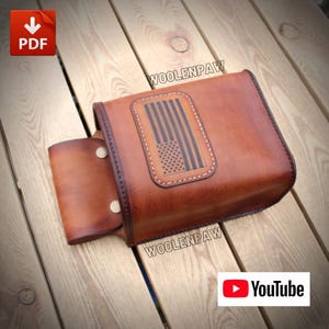 Op de afbeelding: Een bruine leren tas met een rechthoekige patch waarop een gestileerde Amerikaanse vlag is afgebeeld. De tas heeft een klep die wordt vastgezet met twee metalen drukknopen en is afgewerkt met donkerbruine stiksels. De tekst WOOLENPAW is zichtbaar.