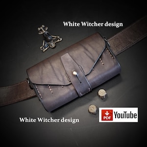 May include: A dark brown leather pouch with a flap closure secured by a single metal stud, featuring visible stitching along the edges. The pouch is attached to a matching leather belt and is accompanied by two metal dice and a metal emblem.