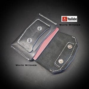May include: A black leather wallet featuring white contrast stitching, two silver-toned metal snap buttons on the front flap, and a second set of snap buttons on the interior. A single pink card is partially inserted. Text reads PDF YouTube WHITE WITCHER.