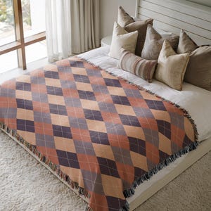 May include: A woven throw blanket featuring a repeating argyle diamond pattern in shades of burnt orange, dark charcoal, and tan. The blanket has a fringed edge and is draped over a bed with white linens and several neutral-toned throw pillows.