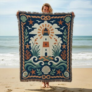 May include: A woven throw blanket featuring a central lighthouse graphic surrounded by a sunburst, clouds, and coastal elements including seashells, starfish, and rope borders in shades of navy blue, cream, sage green, and terracotta orange.