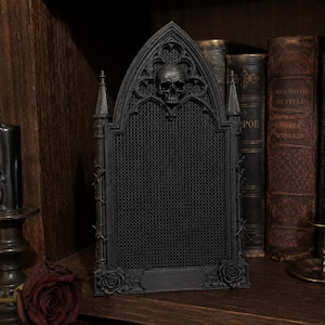 May include: A black, rectangular display board featuring a pointed gothic arch top with a central skull ornament. The board has a fine mesh grid surface, flanked by two small towers with thorny vine details and a rose at each bottom corner.