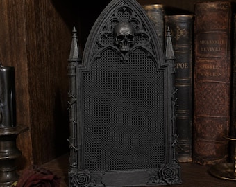 May include: A black, rectangular display board featuring a pointed gothic arch top with a central skull ornament. The board has a fine mesh grid surface, flanked by two small towers with thorny vine details and a rose at each bottom corner.