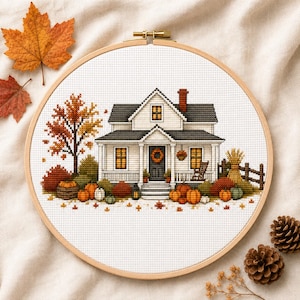 Może przedstawiać: Haft krzyżykowy w okrągłej drewnianej ramce przedstawiający biały dom z ciemnym dachem, kominem i werandą. Scena zawiera drzewo z pomarańczowymi i żółtymi liśćmi, różne dynie, drewniany płot oraz fotel bujany.