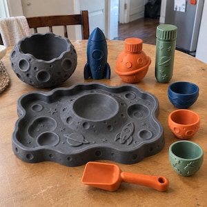Puede incluir: Un juego sensorial con temática espacial que incluye un cuenco y una bandeja de color gris oscuro con cráteres, un cohete azul, un recipiente naranja con forma de planeta, un cilindro verde con motivos espaciales, una pala naranja y tres tazas pequeñas.