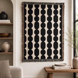 May include: A rectangular woven throw blanket featuring a repeating vertical pattern of solid black elongated oval shapes against a cream-colored background. The blanket has a decorative fringe edge with multi-colored threads along all four sides.