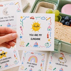 Puede incluir: Una tarjeta cuadrada blanca con una ilustración de un sol amarillo con cara sonriente y el texto YOU ARE MY SUNSHINE, rodeada por un borde colorido de pequeños dibujos que incluyen coches, lápices de colores, estrellas, casas, veleros y manzanas.