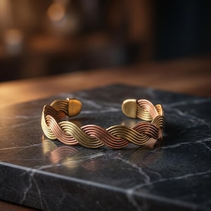 Puede incluir: Una pulsera tipo brazalete construida con múltiples hilos de metal entrelazados en tonos alternos de oro, cobre y bronce. El diseño presenta un patrón trenzado continuo que forma una figura circular con la parte posterior abierta y remates metálicos redondeados.