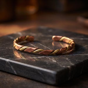 Op de afbeelding: Een armband met een gedraaid ontwerp gemaakt van afwisselende strengen koper en messing. De armband heeft een open achterkant en een gepolijste afwerking, waarbij de roodbruine tint van het koper en de goudgele kleur van het messing zichtbaar zijn.