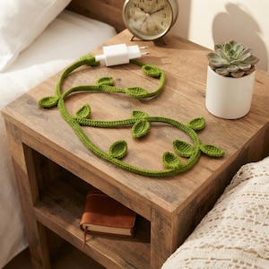 May include: A green crocheted vine cable cover featuring small leaf-shaped accents along its length, attached to a white rectangular power adapter. The cable is coiled in a decorative pattern, showcasing the textured yarn construction and vibrant green color.