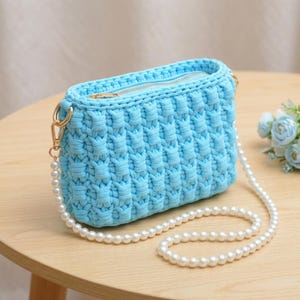 May include: A light blue rectangular crochet handbag featuring a textured woven pattern and a top zip closure. The bag includes a detachable shoulder strap made of white faux pearls attached to the body with gold-toned metal lobster clasps.