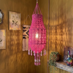 May include: A vibrant hot pink macrame pendant light fixture featuring an intricate knotted cord pattern, a central light bulb, and a series of dangling pink and clear crystal-like beads hanging from the bottom edge of the cylindrical shade.