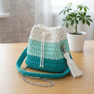 May include: A crochet bucket bag featuring a horizontal color-blocked design in white, light mint green, and dark teal. The bag includes a white drawstring closure, a matching crochet shoulder strap, a silver metal chain, and a white tassel accent.