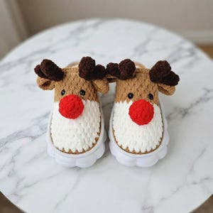 May include: A pair of crochet slippers designed as reindeer, featuring tan yarn tops, white yarn lower sections, dark brown yarn antlers, and bright red yarn noses. The slippers are set on thick, white, rounded rubber-like soles with a scalloped edge.