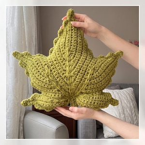 May include: A decorative pillow shaped like a large maple leaf, crafted from olive green yarn with a textured crochet stitch pattern. The leaf features a prominent central vein and multiple pointed lobes, mimicking the natural form of foliage.