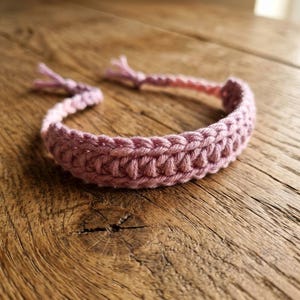 May include: A light pink crochet bracelet featuring a textured, braided stitch pattern. The bracelet is constructed from yarn and includes two loose, thin yarn ties at the ends for fastening, showcasing a consistent, uniform weave throughout the design.
