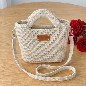 Puede incluir: Un bolso de mano de ganchillo beige con un diseño estructurado, dos asas integradas, una correa de hombro desmontable y pequeños detalles de perlas blancas. Un parche rectangular de cuero marrón en el frente muestra el texto crochet pearl.