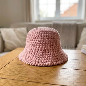 Puede incluir: Un sombrero de pescador de color rosa claro sólido confeccionado con un patrón de ganchillo grueso y texturizado. El sombrero presenta una copa redondeada y un ala inclinada hacia abajo, mostrando una construcción de hilo uniforme.