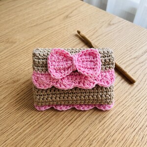 May include: A small rectangular crochet pouch featuring alternating bands of tan and light pink yarn. The front flap is decorated with a prominent pink crocheted bow and a scalloped edge. A wooden crochet hook rests diagonally beside the pouch.