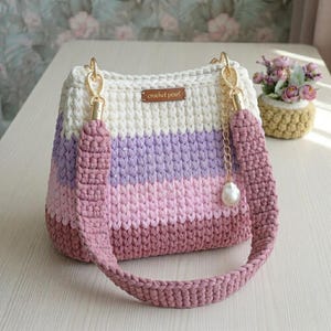 May include: A crochet handbag featuring horizontal stripes in cream, light purple, pink, and mauve. The bag has a thick, matching mauve crochet shoulder strap attached with gold-tone hardware and a small gold chain charm with a white pearl bead.