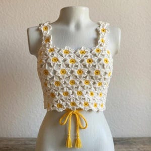 Può includere: Un top corto all'uncinetto senza maniche realizzato con un motivo ripetuto di margherite bianche con centri giallo brillante. Il capo presenta spalline larghe e un cordoncino giallo sull'orlo inferiore con due nappe pendenti.