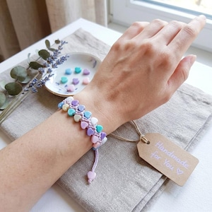 May include: A bracelet featuring small, pastel-colored heart-shaped beads in shades of pink, blue, and purple, strung on a light purple cord. A brown rectangular tag attached to the bracelet displays the text Handmade for You with a heart symbol.