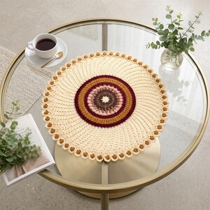 Pode incluir: Um centro de mesa redondo em crochê na cor creme, apresentando um padrão central tipo mandala com anéis concêntricos em tons de castanho escuro, castanho claro e bordeaux profundo. A borda externa tem um acabamento festonado com pequenas bolinhas castanhas em relevo.