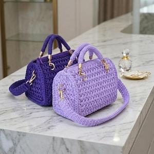 Könnte beinhalten: Zwei tonnenförmige Handtaschen aus geflochtener Kordel. Eine Tasche ist dunkelviolett und die andere helllavendel. Beide verfügen über goldfarbene Metallbeschläge, einschließlich runder Griffbefestigungen und Clips für einen abnehmbaren Schulterriemen.