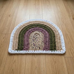 May include: A semi-circular crochet rug featuring concentric bands of color. The design includes layers of white, beige, olive green, and dusty purple yarn, with a speckled beige center. The entire piece is finished with a white crocheted border.
