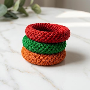 May include: A stack of three circular hair scrunchies made of crocheted yarn. The top scrunchie is bright red, the middle one is vibrant green, and the bottom scrunchie is a warm orange. Each piece features a consistent, textured, woven pattern.
