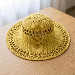 Puede incluir: Un sombrero de ganchillo color amarillo mostaza con una copa redondeada y un ala ancha. El diseño incorpora intrincados patrones calados con recortes repetitivos en forma de diamante alrededor de la base de la copa y el ala.