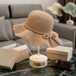 May include: A beige woven straw sun hat with a wide, wavy brim and a decorative bow detail on the side. The hat is displayed on a white stand with a gold-toned rectangular plaque, positioned near two light-colored boxes and dark sunglasses.