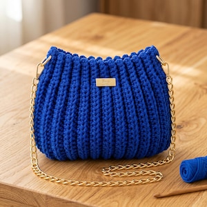 May include: A vibrant royal blue crochet handbag featuring a textured ribbed pattern and a gold-toned metal chain strap. A small rectangular gold-toned metal tag with the text Emy is attached to the front center of the bag.