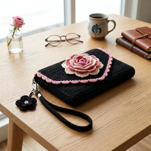 May include: A black rectangular crochet clutch with a wrist strap and a large, layered pink and cream crocheted rose embellishment on the flap. The flap edge features a scalloped pink trim, and a small black flower charm hangs from the wrist strap.