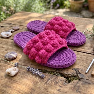 Op de afbeelding: Een paar handgehaakte slippers met een paarse zool en een felroze bovenband met een textuur van nopjessteek. Het open schoeisel is gemaakt van dik garen en toont een duidelijk contrast tussen de dieppaarse basis en het felroze bovengedeelte.