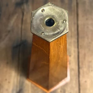May include: A hexagonal wooden kaleidoscope with a brass eyepiece plate featuring three small rivets and an engraved inscription. The body is made of polished light brown wood with a tapered shape, showing a circular dark glass lens at the center.