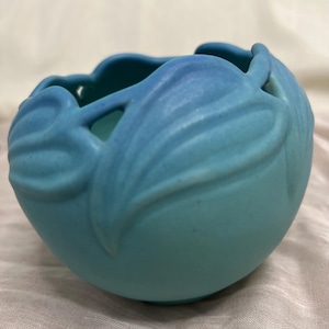 May include: A matte light blue ceramic bowl featuring a raised, sculpted leaf pattern around the rim. The bowl has a rounded, spherical shape with a smooth, uniform finish and a scalloped edge created by the overlapping leaf design elements.