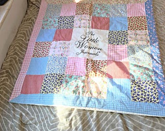 Op de afbeelding: Een vierkante patchwork quilt met diverse stoffen vierkanten in bloemen-, ruit- en streepmotieven in roze, blauwe en witte tinten. Het middelste vierkant is wit met de zwarte tekst The Little Women Journals erop gedrukt.