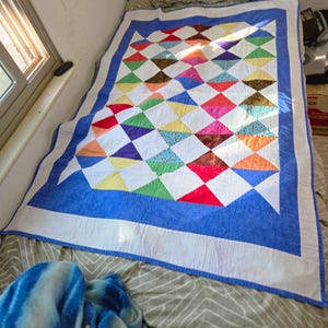 May include: A rectangular quilt featuring a patchwork pattern of multi-colored triangles in shades of red, blue, green, yellow, orange, and purple, interspersed with white triangles. The quilt is bordered by a white band and a blue frame.