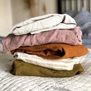 May include: A stack of five folded fabric textiles featuring a variety of colors and textures. The layers include a light beige, a dusty rose, a solid burnt orange, a cream and beige striped pattern, and a solid olive green fabric piece.