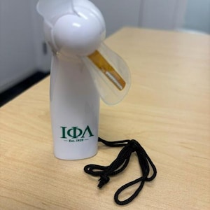 Pode incluir: Um ventilador portátil branco com pás translúcidas e um cordão preto. O corpo do ventilador apresenta as letras gregas verdes IΦΛ e o texto Est. 1929 impresso na frente. O dispositivo tem um topo arredondado e uma base cilíndrica.