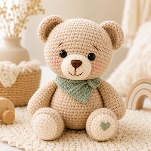May include: A beige crocheted teddy bear featuring a light cream muzzle, dark brown stitched nose, and black safety eyes. The bear wears a sage green triangular neck scarf and has a small green heart embroidered on its left foot pad.