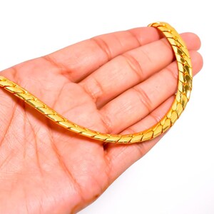 Pode incluir: Um colar de corrente de ouro polido espesso com um design de elos planos entrelaçados. A corrente tem um acabamento metálico brilhante e reflexivo com uma textura suave e contínua em todo o seu comprimento, criando uma aparência ousada.